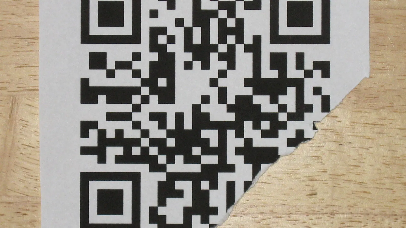 A printed QR code on paper with its lower-right corner torn away, lying on a wooden surface